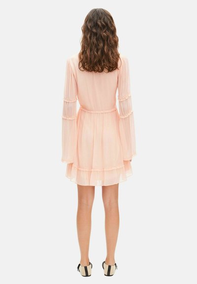 Mujer con cabello castaño ondulado vistiendo un vestido de gasa rosa claro, de manga larga y con volantes, y zapatos planos en blanco y negro, de espaldas.