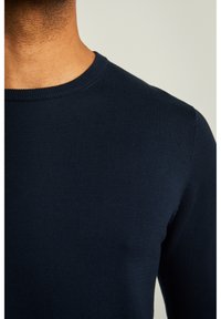 Marineblauer, gerippter Pullover mit Rundhalsausschnitt. Der Stoff wirkt glatt und eng anliegend und zeigt eine Nahaufnahme des oberen Oberkörpers.