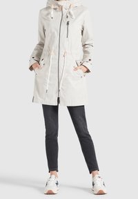 Parka leggera beige con zip frontale, cappuccio regolabile, coulisse in vita e dettagli con bottoni a pressione. Abbinata a jeans scuri e sneaker.
