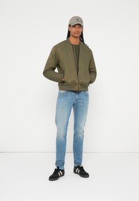 Grøn quiltet bomberjakke med rund hals, parret med lyseblå jeans og sorte sneakers med hvide striber. Grå baseballkasket.
