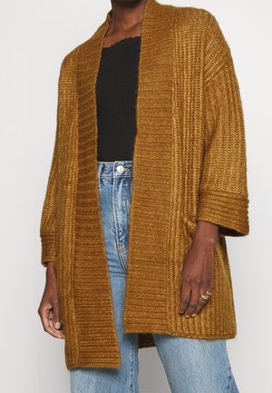 Strickvest in goldbraun mit geripptem Muster und offener Vorderseite. Präsentiert sich mit weiten Ärmeln und einer entspannten Passform, kombiniert mit hoch sitzenden Jeans.