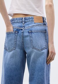 Ανοιχτό μπλε τζιν παντελόνι με ψηλή μέση, με δερμάτινο λογότυπο μάρκας, δύο πίσω τσέπες και άνετη γραμμή. Ορατή τελειωτική ραφή.