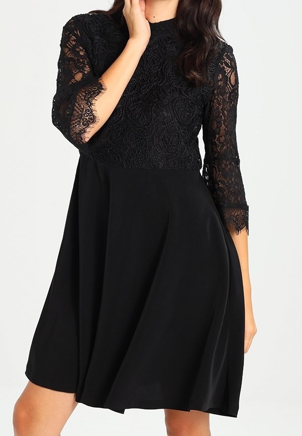 Robe noire avec un corsage en dentelle à motifs floraux, manches trois-quarts, et une jupe fluide en tissu léger.