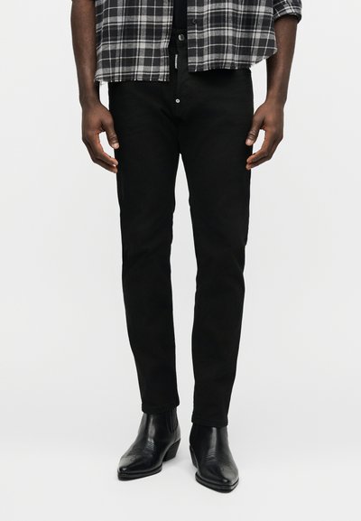 Άνδρας που φοράει μαύρο παντελόνι στενής γραμμής, μαύρα δερμάτινα μποτάκια και ασπρόμαυρο καρό πουκάμισο με μανίκια γυρισμένα.