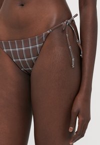 Rjavi karo bikini hlače s vezanjem na boku, ki imajo bele mrežne vzorce in gladko teksturo. Prilagodljiva vezanja za ustrezno prileganje.