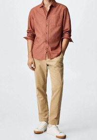 Punainen, teksturoidulla pinnalla varustettu napitettava paita, jossa on rintatasku, yhdistettynä beigeihin housuihin ja valkoisiin lenkkareihin, seisten vaaleanharmaata taustaa vasten.