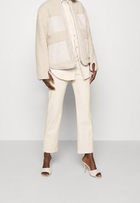 Giacca in shearling beige con toppe in tessuto a contrasto, camicia bianca con bottoni, pantaloni sartoriali avorio e mule con tacco alto aperti leggeri.