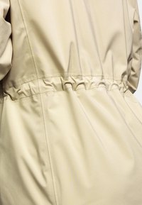 Beige, leichte Jacke mit gerafftem elastischen Bund, glatter Textur und dezentem Glanz. Das Design umfasst gestickte Nähte und einen lässigen Schnitt.