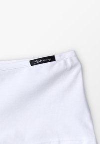 Bela bombažna majica brez rokavov z gladko teksturo. Ima črn napis z logotipom "Skiny" na zgornjem robu. Preprost dizajn, brez vzorcev.