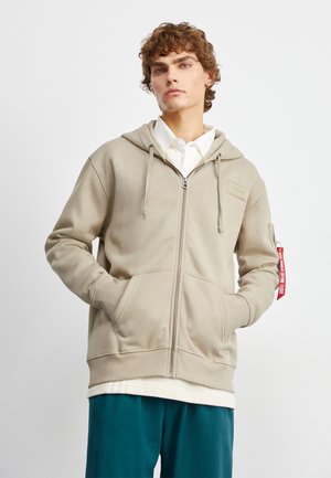 Beige zip-up hoodie met een capuchon, voorzakken, geribde manchetten en een rode tag op de mouw. Gedragen over een wit shirt en tealbroek.