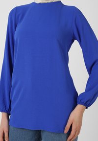 Maglietta a maniche lunghe di un vivace blu, con scollatura rotonda, polsini arricciati e una texture liscia. Abbinata a denim blu chiaro.