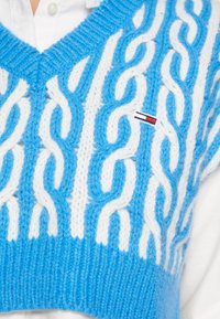 Blå och vit stickad väst med kabelstickning, v-ringning och en liten logotyp med röd, vit och blå rand.