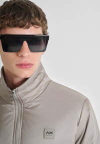 Svijetlosiva puffer jakna s visokim ovratnikom i patentnim zatvaračem; gumeni logotip na prsima. Model nosi crne oversize pravokutne sunčane naočale.