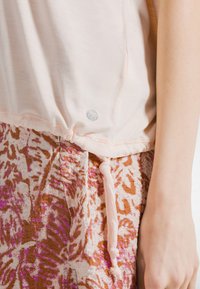 Lyserød top lavet af blødt stof med en rynket kant og sølvlogo. Kombineret med mønstrede, texturerede bukser i brune og lyserøde nuancer.