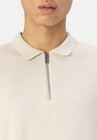 Maglione beige con zip e colletto piatto, realizzato in tessuto testurizzato. La zip in metallo si estende sul davanti, offrendo un tocco funzionale.