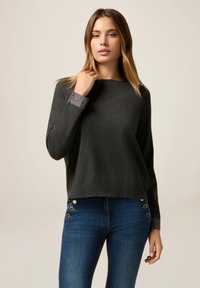 Grå tröja med rund halsringning och långa ärmar, med en texturerad, broderad manschett. Kombinerad med blå smala jeans med gulddetaljer.