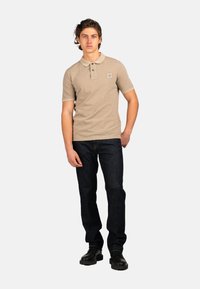 Polo de color beige con un cuello en contraste, mangas cortas y un parche de logo; combinado con unos vaqueros azul oscuro y zapatos negros.