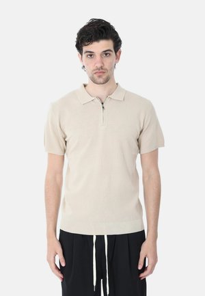 Uomo con capelli scuri e ricci che indossa una polo beige a maniche corte in maglia e pantaloni neri con coulisse bianche, in piedi contro uno sfondo bianco.