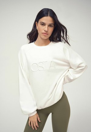Ung kvinde med langt mørkt hår iført hvid OSIA sweatshirt og olivengrønne leggings, poserer med den ene hånd på hoften mod en ensfarvet baggrund.