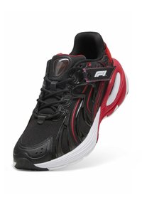 Chaussure de sport noire avec une tige en mesh, des accents rouges et des superpositions noires brillantes. Dispose d'une semelle blanche et d'étiquettes de marque.