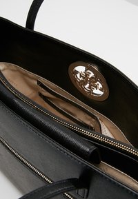 Czarna syntetyczna torebka o gładkiej teksturze, z zamknięciem na zamek i złotym logo. Wnętrze wykończone beżową podszewką i wyposażone w wiele kieszeni.