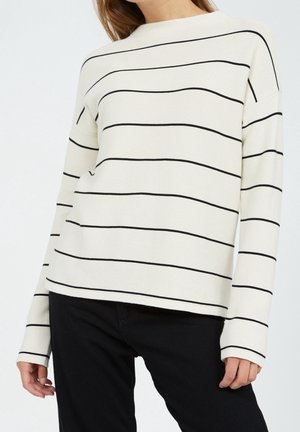 Femme portant un pull à manches longues blanc avec de fines rayures horizontales noires et un pantalon noir, visible du cou aux hanches.