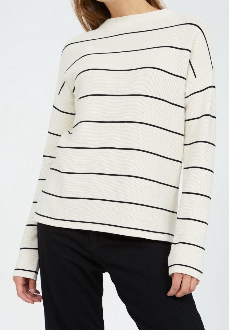 Femme portant un pull à manches longues blanc avec de fines rayures horizontales noires et un pantalon noir, visible du cou aux hanches.