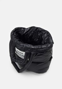 Zwarte gewatteerde tas met twee ronde handvatten, een glanzende textuur en een wit label met "MADS ØRGAAARD COPENHAGEN" aan de zijkant.