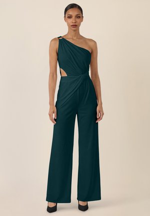 Frau trägt einen dunkeltealfarbenen, einseitigen Jumpsuit mit Cut-out-Detail an der Taille, weitem Bein und schwarzen Absätzen vor einem beigen Hintergrund.