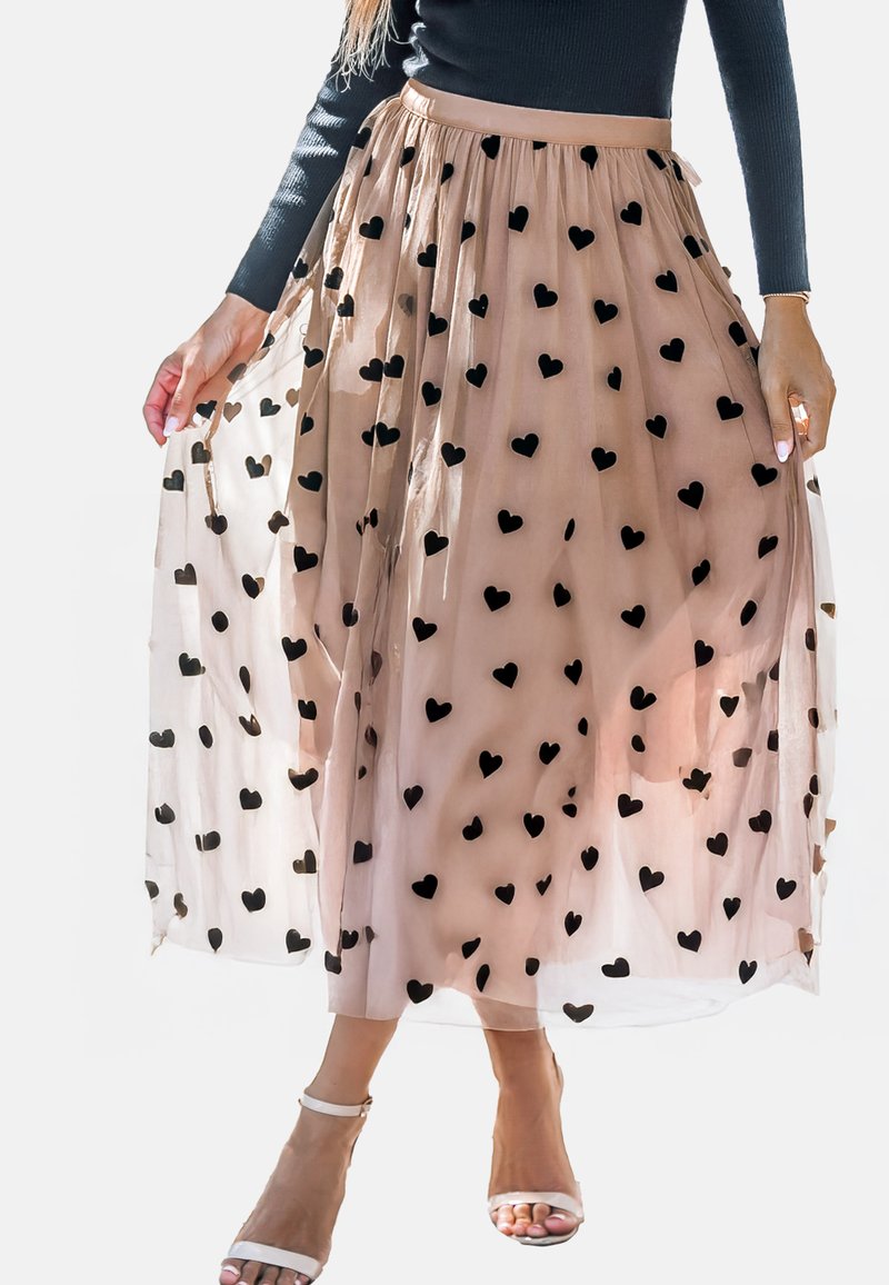 Een beige tule rok met zwarte hartpatronen, met een aansluitende tailleband en een vloeiende silhouette, gecombineerd met witte sandalen met banden.