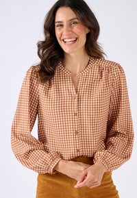 Blusa a scacchi marrone e bianca con collo rotondo, chiusura a bottoni e maniche a sbuffo. Il tessuto sembra leggero, abbinato a una morbida gonna dorata.
