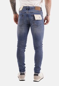 Uomo che indossa jeans blu slim-fit con etichette sulle tasche posteriori, sneakers bianche e una camicia bianca, in piedi contro uno sfondo bianco.