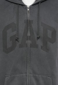 Dark gray zip-up hoodie with drawstrings and large black "GAP" logo printed across the chest above front pockets.