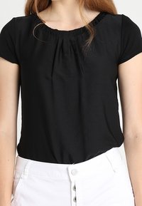 Kobieta w czarnej bluzce z krótkim rękawem i marszczonym dekoltem oraz białych spodniach z wysokim stanem i zapięciem na guzik.