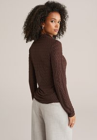 Rjav top z dolgimi rokavi z teksturiranim vzorcem. Prilegajoč kroj, z okroglim izrezom in rahlo nabrano zadnjo stranjo. Kombiniran s svetlo sivimi hlačami.