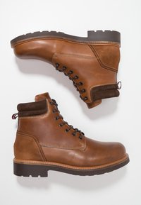 Hnědé kožené kotníkové boty s kulatou špičkou, tmavohnědými akcenty, šněrováním, texturovaným svrškem a gumovou podrážkou s vzorovaným dezénem.