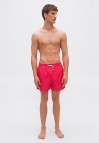 Νέος άνδρας στέκεται ξυπόλητος φορώντας φωτεινό ροζ μαγιό με λευκό κορδόνι, κοιτάζοντας προς το πλάι με φόντο έναν απλό ανοιχτόχρωμο τοίχο.