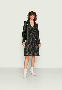 Jaqueta de motociclista de couro preto sobre um vestido preto estampado com designs geométricos. Modelo usa botas de tornozelo claras. Texturas suaves e cores contrastantes.
