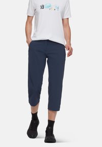 Mörkblå korta byxor i lätt material, med en avslappnad passform, framfickor och en diskret logotyp på sidan. Bar med en vit t-shirt och svarta skor.