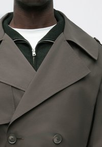 Double-breasted gray coat with broad lapels, featuring two buttons, layered over a green ribbed zip-up sweater. Smooth fabric texture.