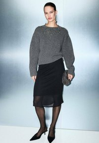 Suéter de punto gris con cuello texturizado, falda asimétrica negra, medias negras transparentes y zapatos negros de punta. Sosteniendo un bolso de mano de cuentas.