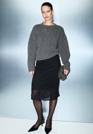 Suéter de punto gris con cuello texturizado, falda asimétrica negra, medias negras transparentes y zapatos negros de punta. Sosteniendo un bolso de mano de cuentas.