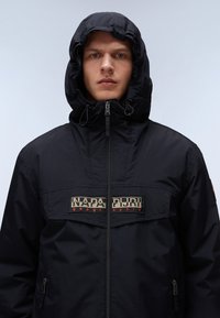 Schwarze gepolsterte Jacke mit einem Reißverschluss vorne, verstellbare Kapuze und einem Logo-Patch auf der Brust. Besitzt eine glatte Textur und ein praktisches Design.