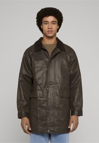 Brun skinnjakke med teksturert finish, har en manchesterkrage, to frontlommer og knappelukking med trykknapper, brukt over en grønn skjorte.