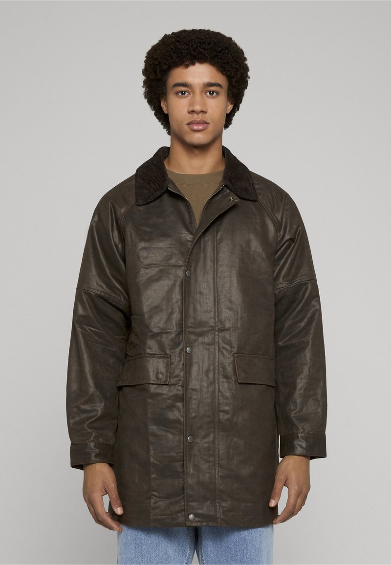 Brun skinnjakke med teksturert finish, har en manchesterkrage, to frontlommer og knappelukking med trykknapper, brukt over en grønn skjorte.