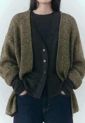 Femme portant des cardigans marron et noir superposés et un jean bleu foncé, les mains dans les poches, sur un fond uni.