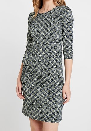 Femme portant une robe ajustée mi-longue avec des manches trois-quarts et un motif géométrique bleu marine sur un fond clair.