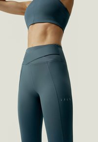 Höga, mörk teal leggings med en slät yta och design av korsettsmidda midja. Har subtil logotyp på underbenet.