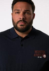 Námořnické polo tričko s krátkým rukávem, třemi knoflíky, límečkem a červeně vyšívaným logem "MOUNTAIN ADVENTURE" na levé straně hrudi.