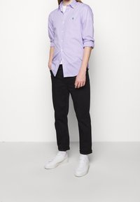 Man som bär en ljuslila randig knäppskjorta, svarta byxor, vita strumpor och vita sneakers, stående mot en vit bakgrund.